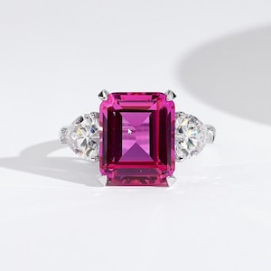Peut inclure: Une bague en argent ornée d'une grande pierre précieuse magenta rectangulaire au centre, flanquée de deux pierres précieuses claires en forme de poire. La bague est posée sur un fond blanc.
