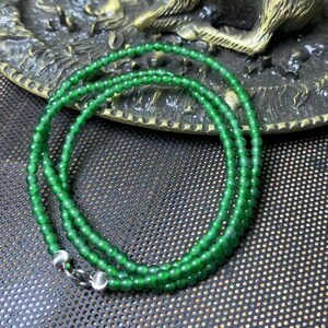 May include: A vibrant green beaded necklace with a silver clasp. The necklace is composed of small, round beads, creating a delicate and elegant appearance. The beads are a rich, emerald green color, and the necklace is displayed on a textured surface.
