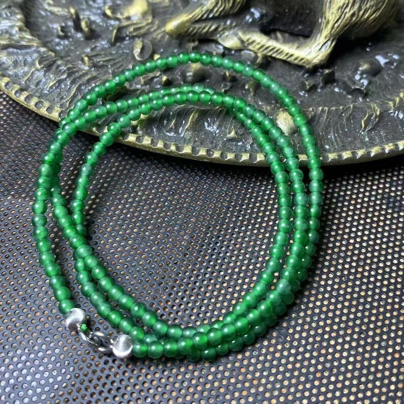 May include: A vibrant green beaded necklace with a silver clasp. The necklace is composed of small, round beads, creating a delicate and elegant appearance. The beads are a rich, emerald green color, and the necklace is displayed on a textured surface.