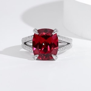 Puede incluir: Un anillo de plata con una gran gema roja oscura tallada en cojín. La banda se divide en dos filas, cada una incrustada con pequeñas gemas transparentes. El diseño del anillo es elegante y llamativo, adecuado para ocasiones especiales.