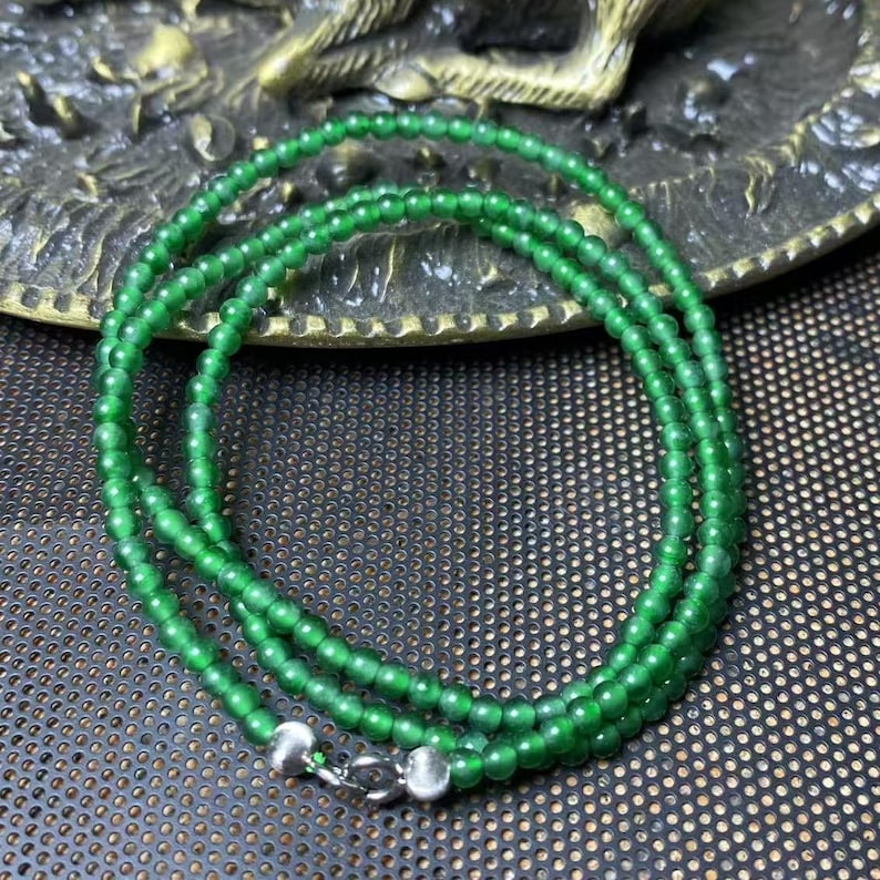 May include: A vibrant green beaded necklace with a silver clasp. The necklace is composed of small, round beads, creating a layered effect. The beads have a glossy finish, reflecting light. The necklace is displayed on a textured surface.