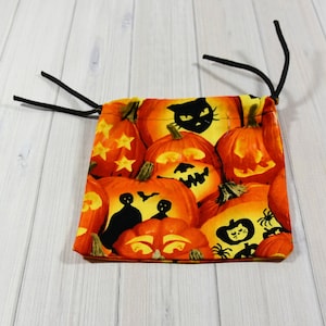 Op de afbeelding: Een kleine, vierkante trekkoordtas met een Halloween-patroon. De stof heeft een herhalend ontwerp van oranje pompoenen met uitgesneden gezichten, zwarte katten en spinnen. De tas heeft een zwarte trekkoordsluiting.