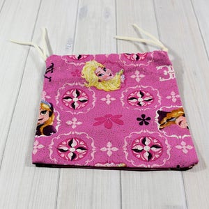 May include: A pink fabric bag featuring a repeating pattern of a cartoon character and floral designs. The bag has a drawstring closure with white cords. The bag is made of a soft, patterned fabric.