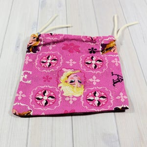 May include: A small, pink drawstring bag featuring a repeating pattern of floral and geometric designs, with an image of a blonde character. The bag has white drawstrings and is likely made of fabric.