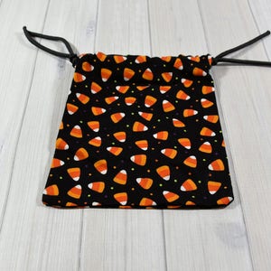 May include: A black drawstring bag patterned with orange, yellow, and white candy corn. The bag is open, with black drawstrings. The background is a light wood surface.