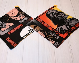 Könnte beinhalten: Zwei quadratische Topflappen im Star Wars-Design. Einer zeigt Han Solo und einen Sturmtruppler, der andere Darth Vader. Die Topflappen sind schwarz mit orangefarbenen, roten und weißen Akzenten. Jeder hat eine gelbe Schlaufe zum Aufhängen.