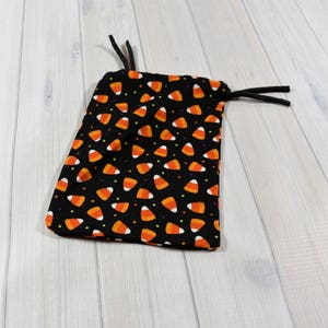 May include: A black drawstring bag patterned with orange, yellow, and white candy corn. The fabric bag has black drawstrings. The bag is on a light wood surface.