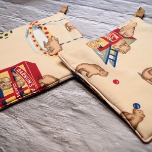 May include: Two square pot holders with a cream-colored background featuring a circus bear print. The design includes circus scenes, bears, and the text "Barnum's Animals Crackers". Each pot holder has a brown loop.