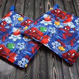 Puede incluir: Dos agarraderas de olla azules y rojas con un tema navideño de invierno con un personaje de Spiderman de dibujos animados. Las agarraderas tienen un patrón de copo de nieve y adorno.