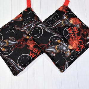 May include: A pair of black and red pot holders with a repeated pattern of a superhero character in a black suit and red hair. The text "BLACK WIDOW" is printed on the pot holders.