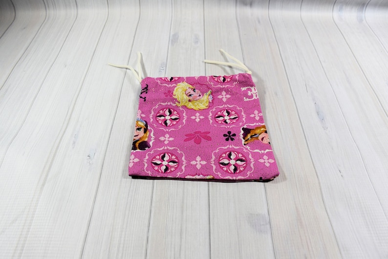May include: A pink fabric bag featuring a repeating pattern of a cartoon character and floral designs. The bag has a drawstring closure with white cords. The bag is made of a soft, patterned fabric.