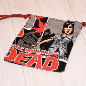 May include: A small drawstring bag featuring a comic book design with the words "THE WALKING DEAD" in red and white. The bag has a gray and black background with a character illustration. The drawstring is a reddish-brown cord.