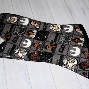 May include: A gray oven mitt featuring a Star Wars theme. The mitt is patterned with various characters, logos, and the words "Star Wars" in a repeating grid. The edges are trimmed with black fabric and a loop for hanging.