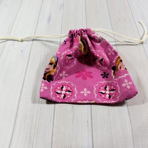May include: A pink drawstring bag featuring a repeating pattern of a character's face, flowers, and geometric shapes. The bag has a white drawstring closure. The bag is made of fabric.
