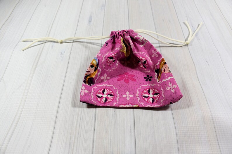 May include: A pink drawstring bag featuring a repeating pattern of a character's face, flowers, and geometric shapes. The bag has a white drawstring closure. The bag is made of fabric.