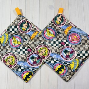 May include: Two brown and white square pot holders with a checkered pattern and images of Wonder Woman, Batgirl, and Superman. The pot holders have yellow loops for hanging.