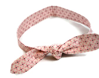May include: A pink and white patterned headband with a knotted bow. The fabric features a repeating pattern of pink leaves and black dots. The headband is designed to be worn around the head.