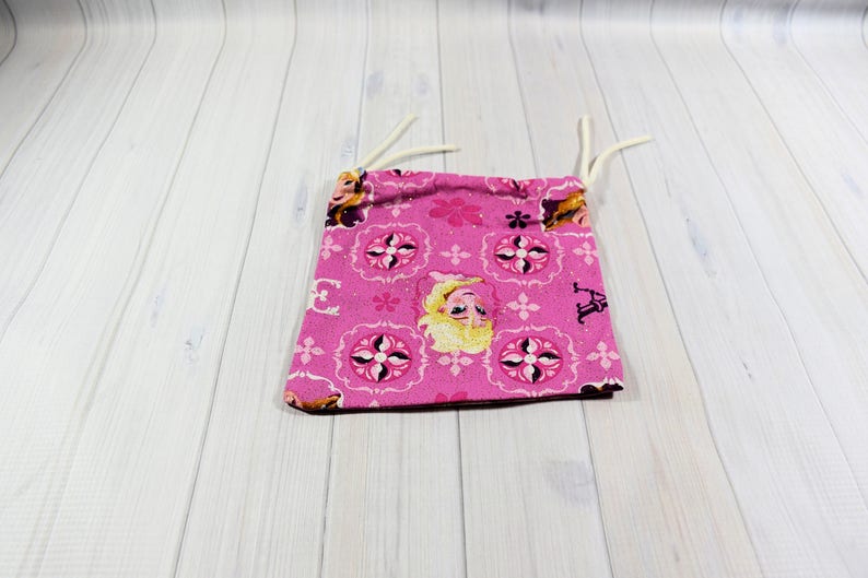 May include: A small, pink drawstring bag featuring a repeating pattern of floral and geometric designs, with an image of a blonde character. The bag has white drawstrings and is likely made of fabric.