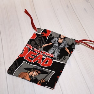 May include: A small drawstring bag featuring a comic book design with the words "THE WALKING DEAD" in red and white. The bag has a black background with images of characters from the series. Red drawstring closure.
