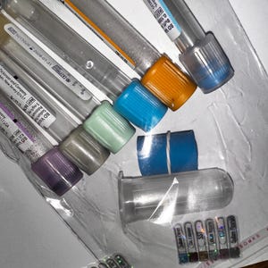 Könnte beinhalten: Eine Sammlung klarer Blutentnahmeröhrchen aus Kunststoff mit verschiedenfarbigen Verschlüssen, darunter orange, blau und lila. Einige Röhrchen haben Etiketten mit Text. Die Röhrchen sind auf einer transparenten Kunststofffolie angeordnet.