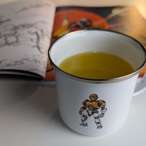 Könnte beinhalten: Eine weiße Emailletasse mit silbernem Rand, gefüllt mit einer gelben Flüssigkeit. Die Tasse zeigt ein Motiv eines Roboters in einem gelben und orangenen Anzug.