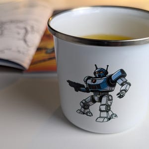 Op de afbeelding: Een witte emaille mok met een blauwe en witte robot illustratie. De mok heeft een zilveren rand en handvat. De robot houdt een pistool vast en heeft een rood oog.