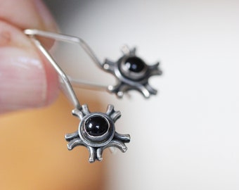 May include: A pair of silver-toned earrings with a unique design. Each earring features a central black stone surrounded by a starburst-like pattern. The earrings are held up by a wire hook.