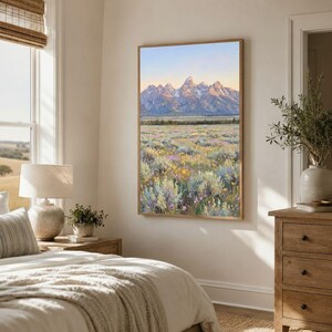 May include: A vertical framed wall art print depicting a landscape scene with a vast field of wildflowers in the foreground and a range of snow-capped mountains under a soft, pale blue and golden sky in the background, mounted on a wall.
