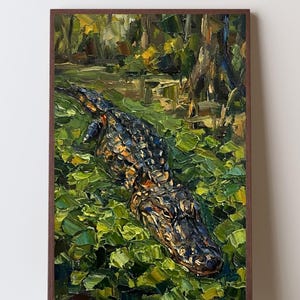 Könnte beinhalten: Ein Ölgemälde in einem braunen Rahmen zeigt einen Alligator, der teilweise in einem mit leuchtend grünen Seerosen bedeckten Sumpf untergetaucht ist. Das Kunstwerk verwendet dicke, strukturierte Pinselstriche für die dunkle, schuppige Haut.