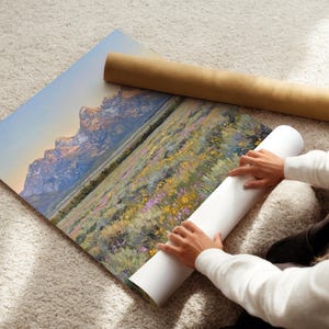 May include: A large rectangular art print featuring a landscape painting of jagged, snow-capped mountains at sunrise, overlooking a field of yellow and purple wildflowers. The print is partially rolled, with a brown cardboard shipping tube nearby.