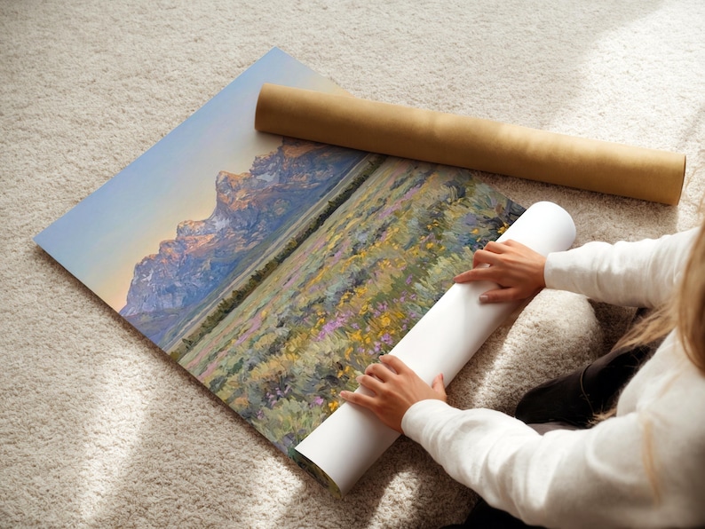 May include: A large rectangular art print featuring a landscape painting of jagged, snow-capped mountains at sunrise, overlooking a field of yellow and purple wildflowers. The print is partially rolled, with a brown cardboard shipping tube nearby.