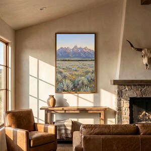 May include: A framed landscape painting depicting a mountain range with snow-capped peaks under a soft sky, overlooking a vast field of sagebrush and wildflowers in shades of green, yellow, and purple, displayed on a light-colored wall.