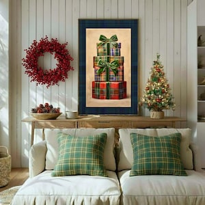 May include: A framed wall art print featuring a stack of three wrapped gift boxes with green bows, displayed above a sofa with two green plaid throw pillows. The artwork is centred on a white panelled wall between a red berry wreath and a small tree.
