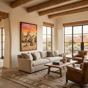 May include: A framed wall art print featuring a painting of two coyotes in a desert landscape with saguaro cacti under a vibrant orange and pink sunset sky. The artwork is displayed on a light-colored wall above a beige sofa in a living room.