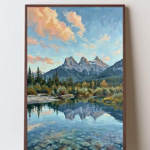 May include: A framed landscape painting depicting three jagged mountain peaks under a blue sky with soft pink and orange clouds. A dense forest of evergreen trees sits below the mountains, reflected in a clear, shallow, rocky river.