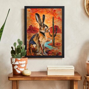 Puede incluir: Una impresi&oacute;n art&iacute;stica enmarcada que presenta una pintura al &oacute;leo de una liebre en un paisaje des&eacute;rtico. La escena incluye un cactus saguaro alto, formaciones rocosas rojas y un arroyo azul bajo un cielo de atardecer naranja y rojo.