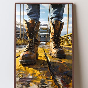 Könnte beinhalten: Ein gerahmter Wandkunstdruck, der ein Paar abgenutzte braune Leder-Arbeitsstiefel mit schwarzen Schnürsenkeln und eine blaue Jeans zeigt. Die Stiefel stehen auf einer gelben Metallbrücke, im Hintergrund ist ein Stadion zu sehen.