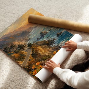 May include: An oil painting of a coastal landscape with a winding road, in shades of orange, blue, and brown. A rolled-up canvas and a brown cardboard tube are next to the painting. The painting is on a white surface.