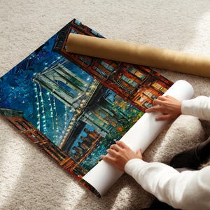 May include: A colorful print of a cityscape, featuring a bridge and buildings, is laid out on a soft, light-colored surface. A rolled-up print and a cardboard tube are next to the print. The print is being handled.