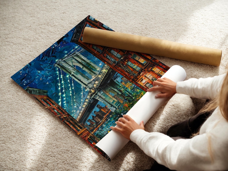 May include: A colorful print of a cityscape, featuring a bridge and buildings, is laid out on a soft, light-colored surface. A rolled-up print and a cardboard tube are next to the print. The print is being handled.