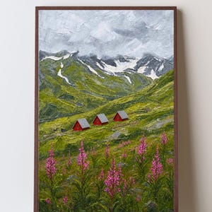 May include: An oil painting in a dark brown frame depicting a mountain landscape with three small red cabins nestled in a green valley. The foreground features tall pink wildflowers, while the background shows snow-capped peaks under a cloudy sky.