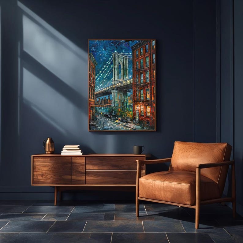 May include: A framed painting of the Brooklyn Bridge at night, with illuminated lights and surrounding buildings. The artwork is displayed above a wooden cabinet and a leather armchair in a room with dark blue walls.