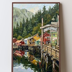 May include: An oil painting in a brown frame depicting Creek Street with colourful wooden houses built on stilts over water. A wooden sign reads Creek Street above a boardwalk, with dense green pine trees and a mountain range in the background.
