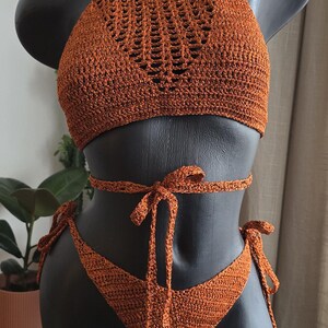 May include: A rust-colored crochet bikini set featuring a high-neck halter top with an intricate open-stitch pattern and a matching bottom with side ties. A long, thin crochet cord wraps around the waist and ties in the front center.