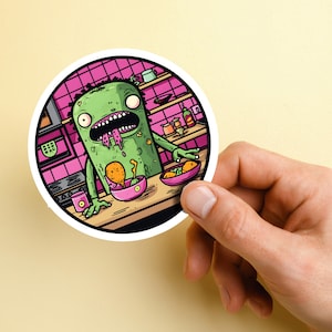 Pode incluir: Um monstro verde com uma língua rosa e uma tigela rosa e branca de comida na sua frente. O monstro está em uma cozinha com azulejos rosa e prateleiras.