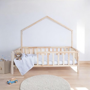 Peut inclure: Un lit en bois pour enfant en bas âge avec une tête de lit en forme de maison et une couverture blanche. Le lit a un matelas blanc et un ours en peluche sur la couverture.