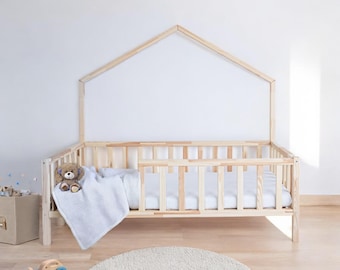 Könnte beinhalten: Ein Kinderbett aus Holz mit einem hausförmigen Kopfteil und einer weißen Decke. Das Bett hat eine weiße Matratze und einen Teddybären auf der Decke.