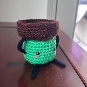 May include: A handmade crochet figure with a brown hat and a green body. The figure has black arms and legs, and a smiling face with pink cheeks. The figure is sitting on a wooden surface.