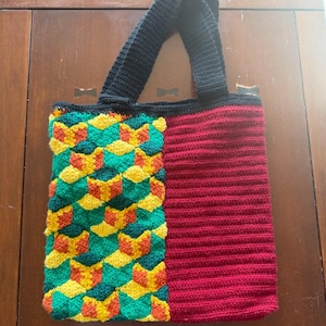 May include: A handmade crochet tote bag with a black handle and trim. The bag features a vibrant design with a geometric pattern in green, yellow, and orange on one side, and a solid red ribbed pattern on the other.
