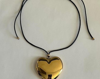 May include: A gold-colored heart pendant necklace with a black cord. The heart is a shiny, reflective gold. The cord is adjustable with small gold-colored beads at the ends.